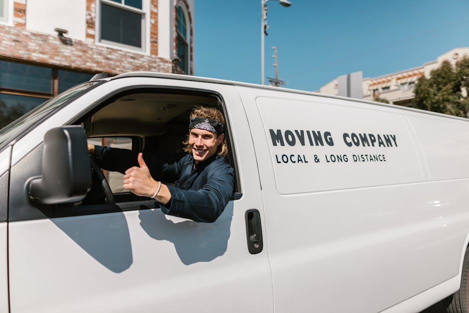 Inside a white moving van parked on a street in Pimlico with a young male mover visible through the open driver's side window. The mover has light brown, curly hair, is wearing a black headband and a 
