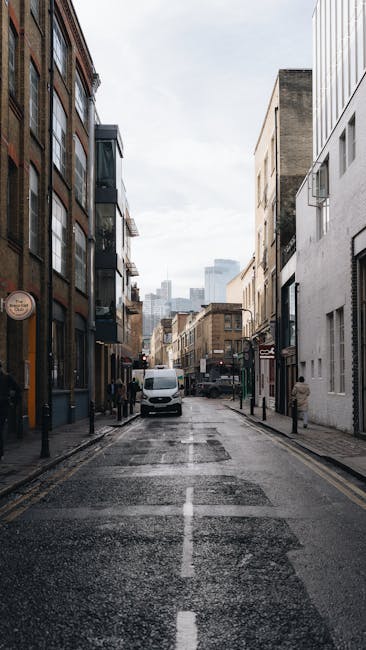 Inside a white moving van parked on a street in Pimlico with a young male mover visible through the open driver's side window. The mover has light brown, curly hair, is wearing a black headband and a 