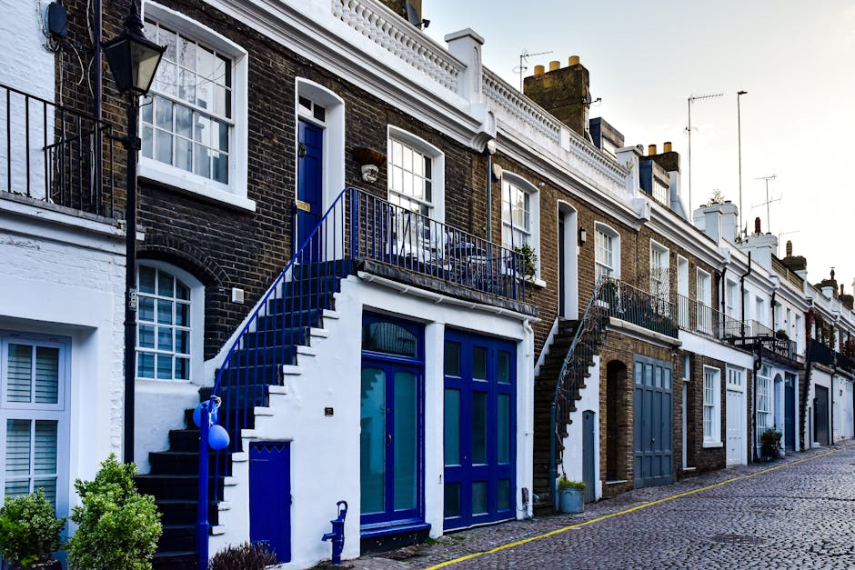 A row of terraced houses on a cobbled street, with each property featuring brick facades and white window frames. The house in the foreground has a distinctive blue front door, a small balcony with a 
