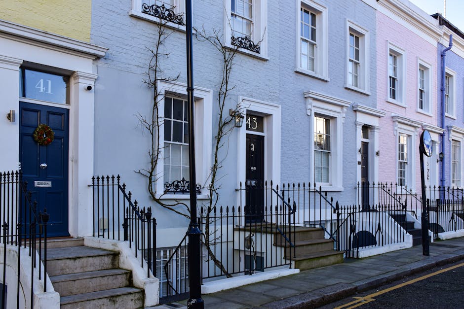 A row of terraced houses on a cobbled street, with each property featuring brick facades and white window frames. The house in the foreground has a distinctive blue front door, a small balcony with a 
