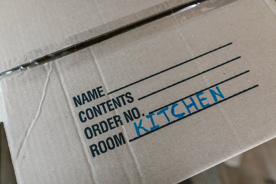 Close-up of a cardboard moving box labeled with black printed text for 'NAME', 'CONTENTS', 'ORDER NO.', and 'ROOM', with the word 'KITCHEN' handwritten in blue ink under the 'ROOM' section. The box appears to be part of a home relocation process, possibly inside a property or near a loading area, prepared for furniture transport and packing during a house move. The cardboard material is standard, and the image emphasizes organized packing for relocation services provided by Storage Pimlico. The background shows additional boxes and the environment suggests an interior space ready for a professional removal company's loading process as part of house removals or moving services.