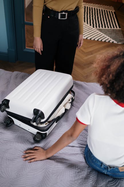 An open glass-front suitcase on a light pink background, filled with travel essentials such as a tube of cream, sunglasses, a small camera, a tablet, a notebook, and assorted clothing items folded and
